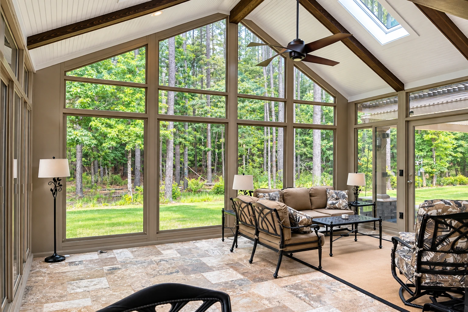 A bright four-season sunroom has tall windows, exposed dark wood beams, a ceiling fan, and seating overlooking trees.