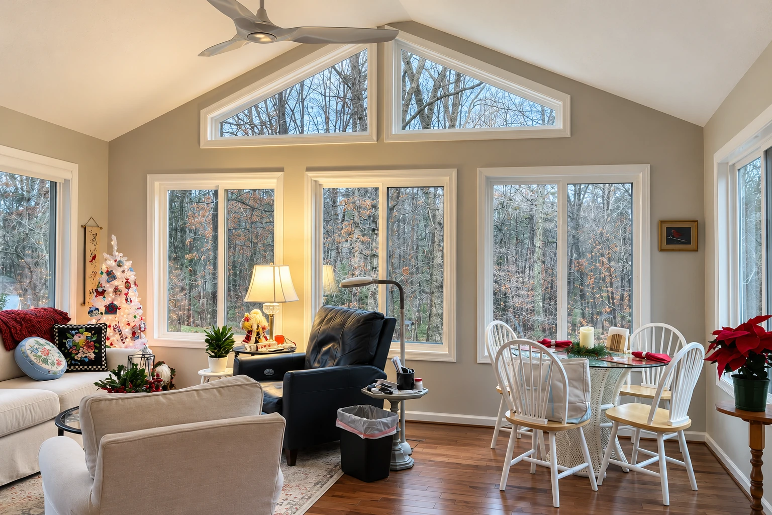 A bright four-season sunroom has tall windows, a vaulted ceiling, sofas, a dining table, and a decorated tree beside a wintry woodland view.