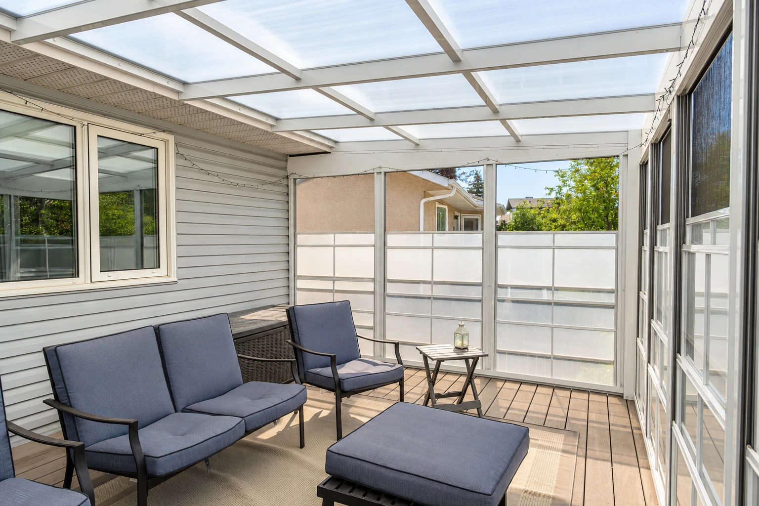A screened room with a translucent paneled roof has gray siding, blue lounge chairs, and half-height privacy panels.