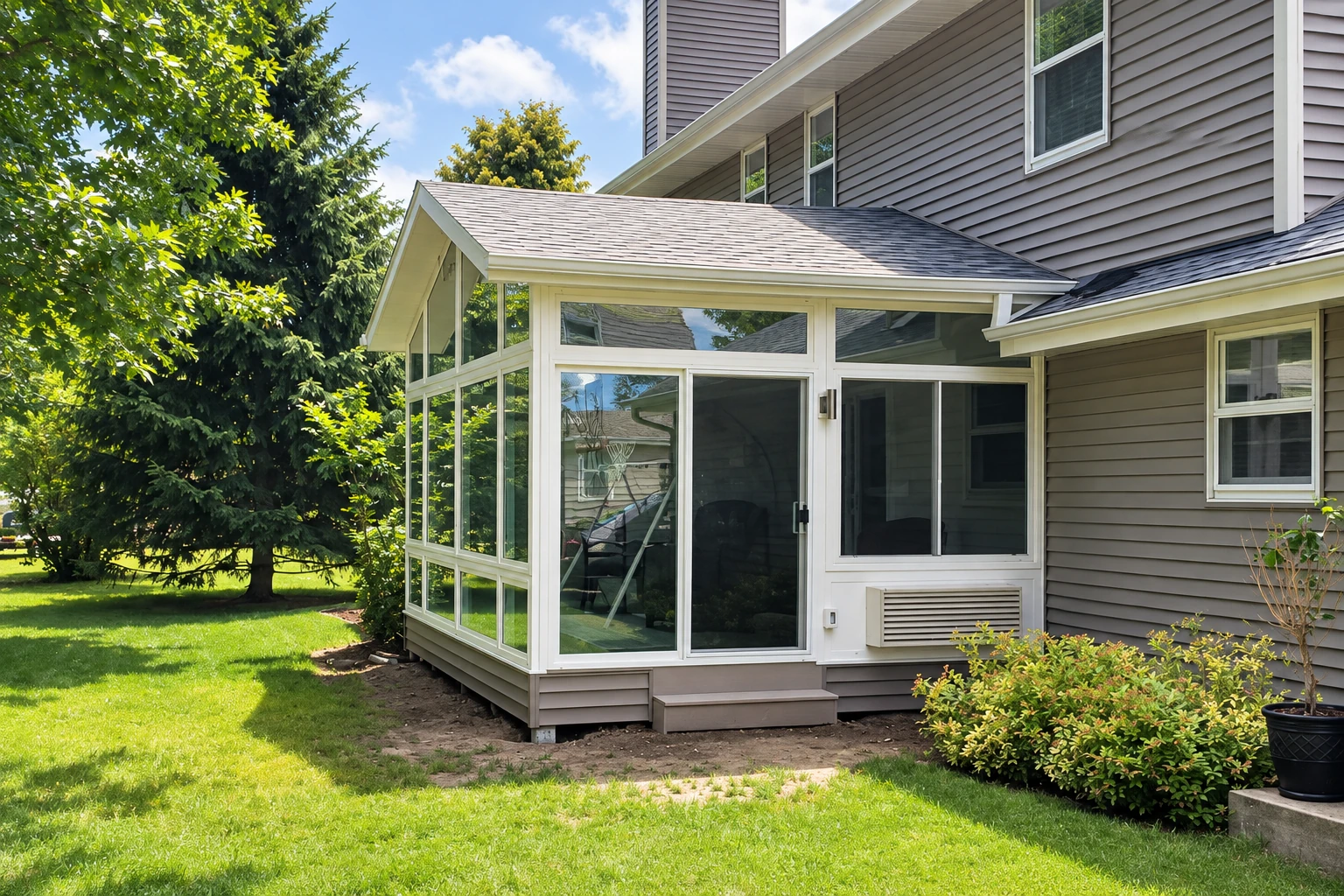 A glass sunroom addition with white frames and a pitched roof extends from a gray-sided house into a leafy backyard.