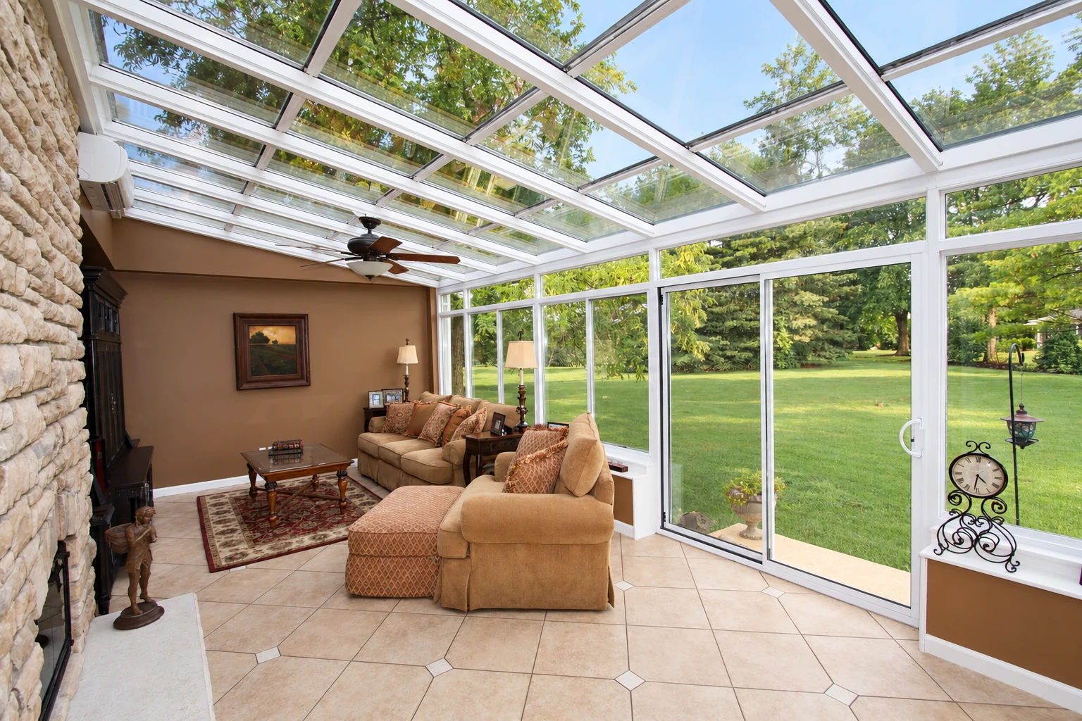 Sunroom with a glass-paneled roof, floor-to-ceiling windows, tiled floor, and tan seating overlooking a lawn.