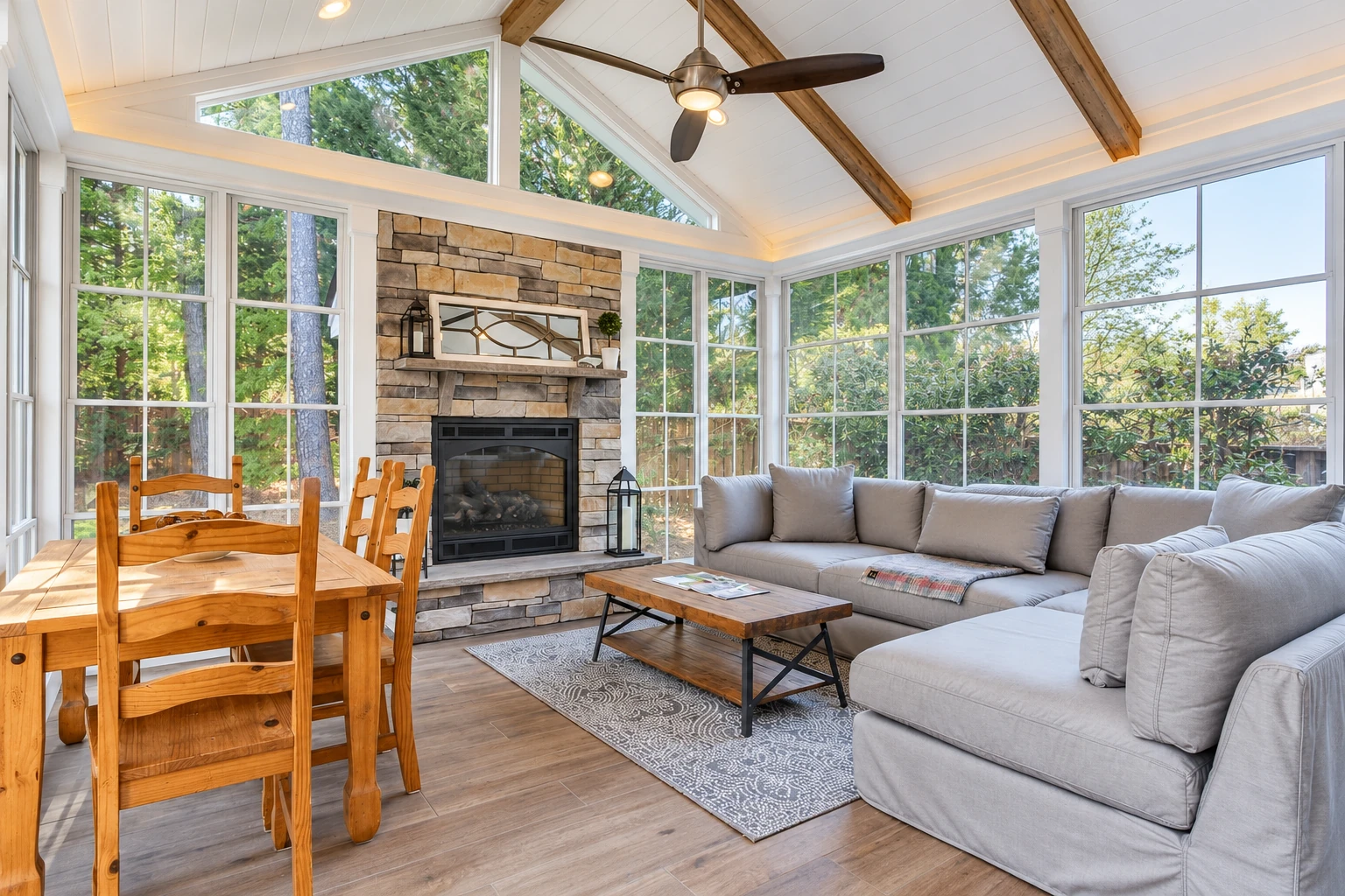 Bright sunroom with a stone fireplace, vaulted wood-beam ceiling, large windows, gray sectional, and wooden dining set.