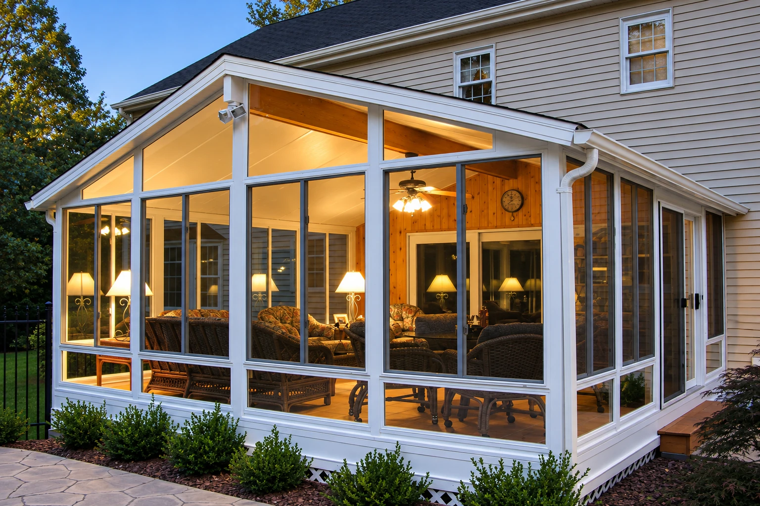 A white-framed, gabled vinyl sunroom extends from a beige-sided house, with large windows and warmly lit wicker seating inside.