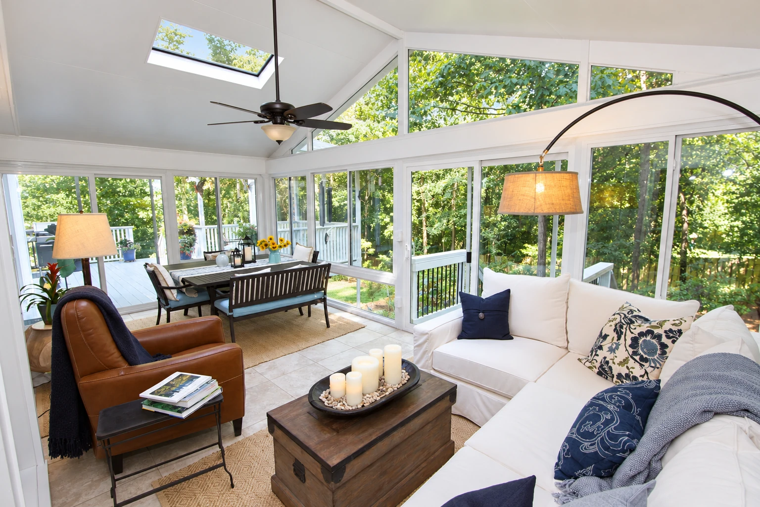 White vinyl sunroom with expansive windows, skylight, dining area, and a white sofa beside a wooded deck.